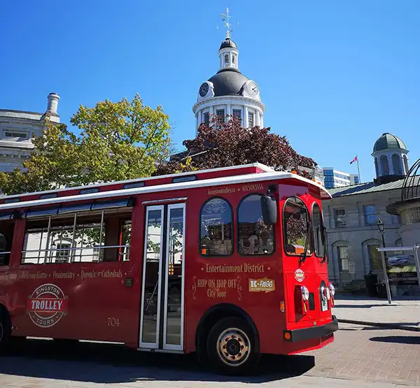 Red trolley driving past Kingston's City Hall, a large, 4 storey limestone building with a domed clock tower in the center.
